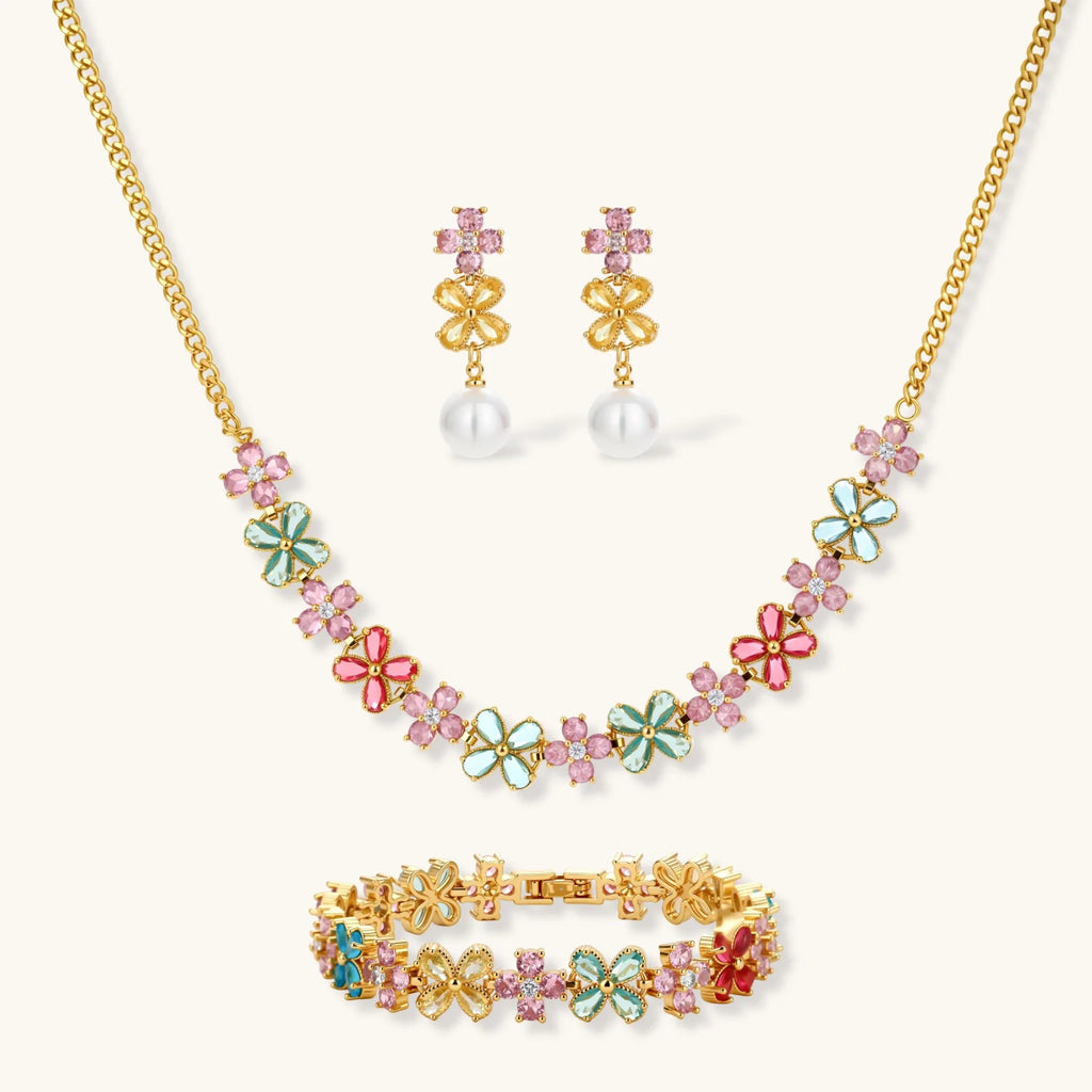 Gold-plated flower necklace, bracelet and drop earrings with multicolor zirconia petals and white round beads on a cream background.
