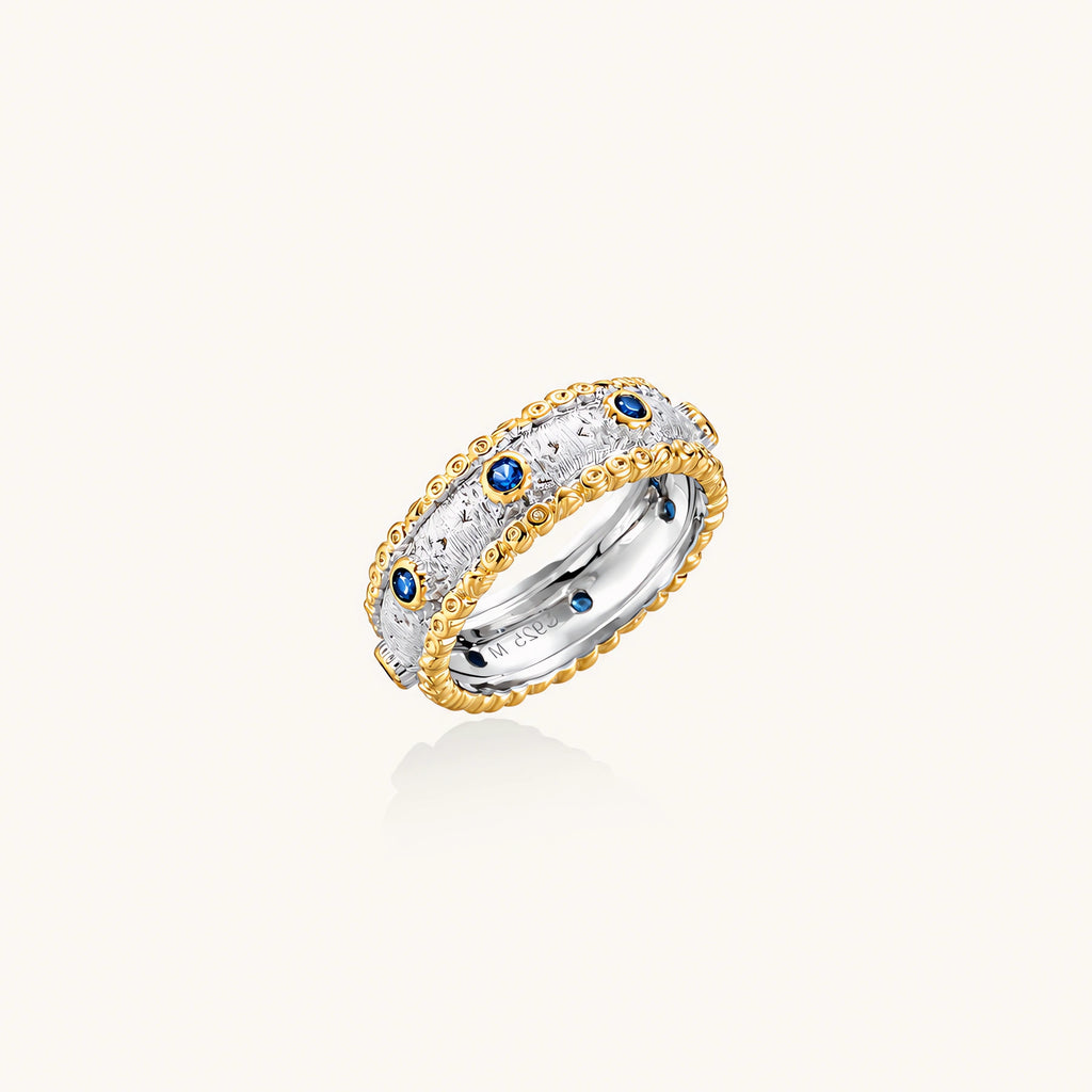 Two-tone ring with brushed silver center, gold beaded edges, and blue zirconia stones on white background.