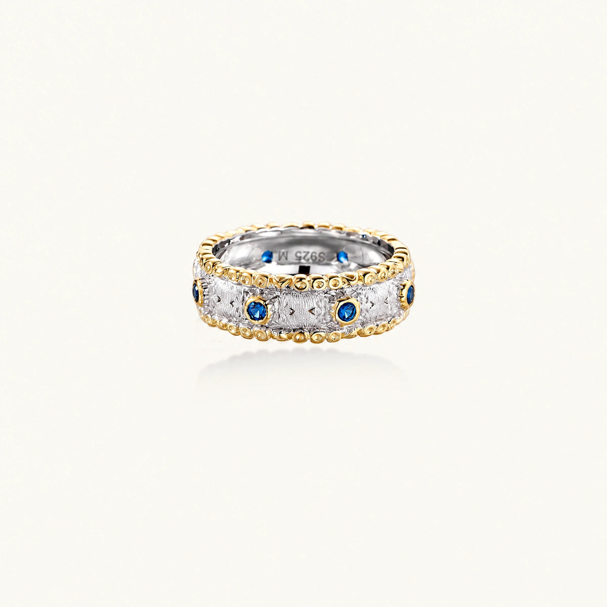 Two-tone ring with brushed silver center, gold beaded edges, and blue zirconia stones on white background.