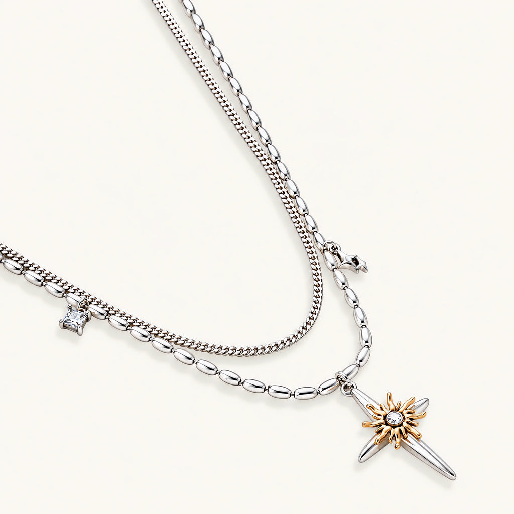 Layered silver-toned necklace set with cross pendant and gold sun charm detail, matching drop earrings on white background.