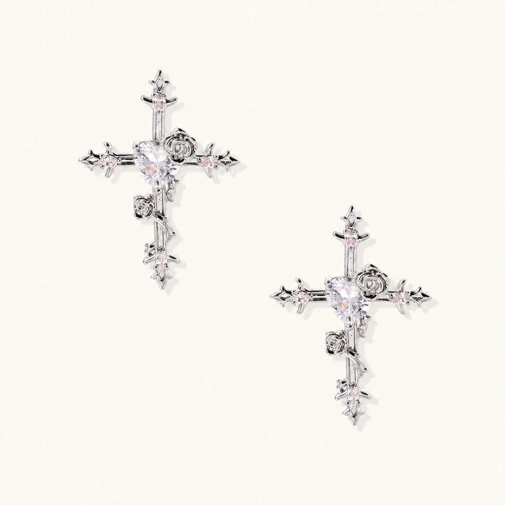 Pair of ornate silver cross earrings with heart-cut clear zirconia center, rose motifs, and spike tips on cream background.