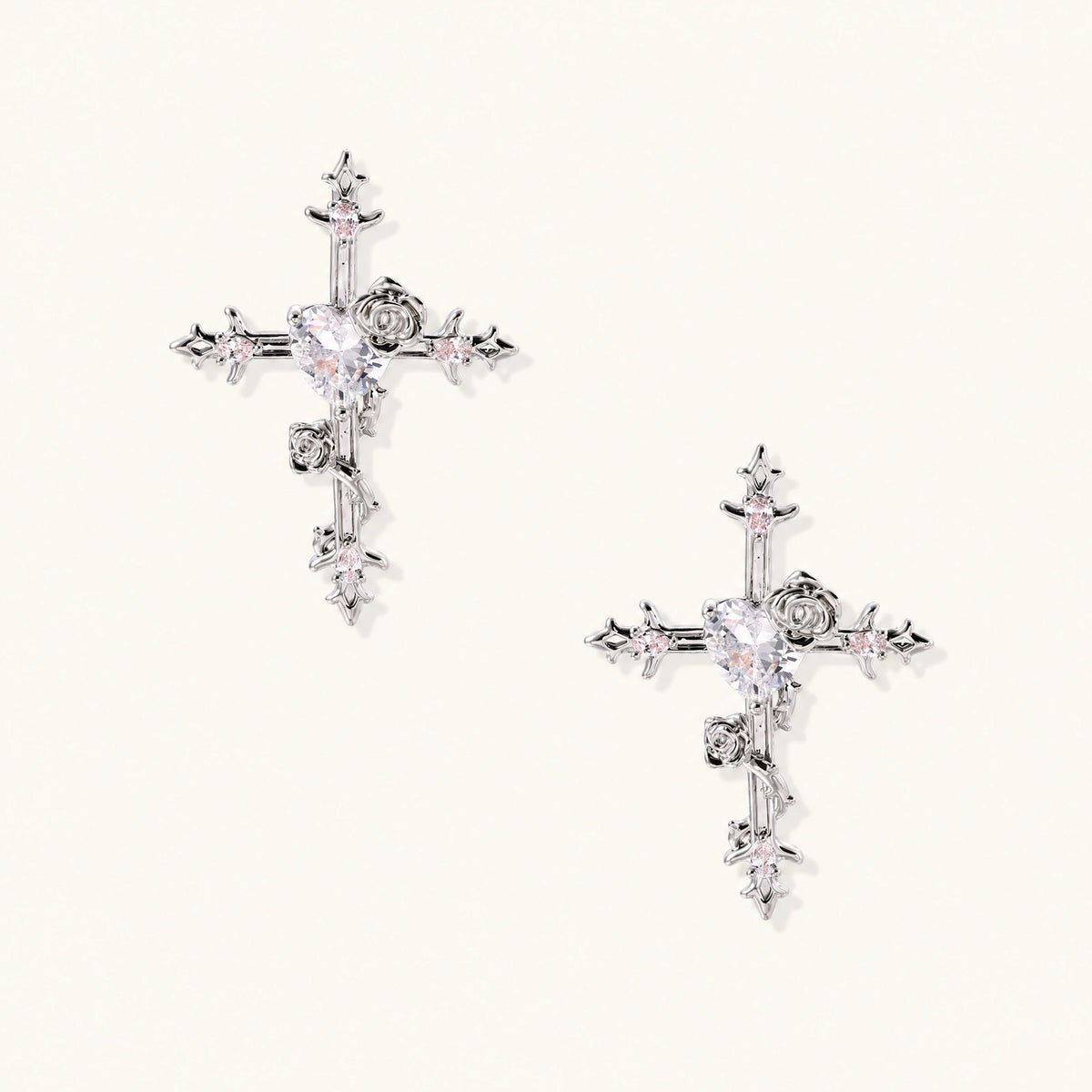 Pair of ornate silver cross earrings with heart-cut clear zirconia center, rose motifs, and spike tips on cream background.